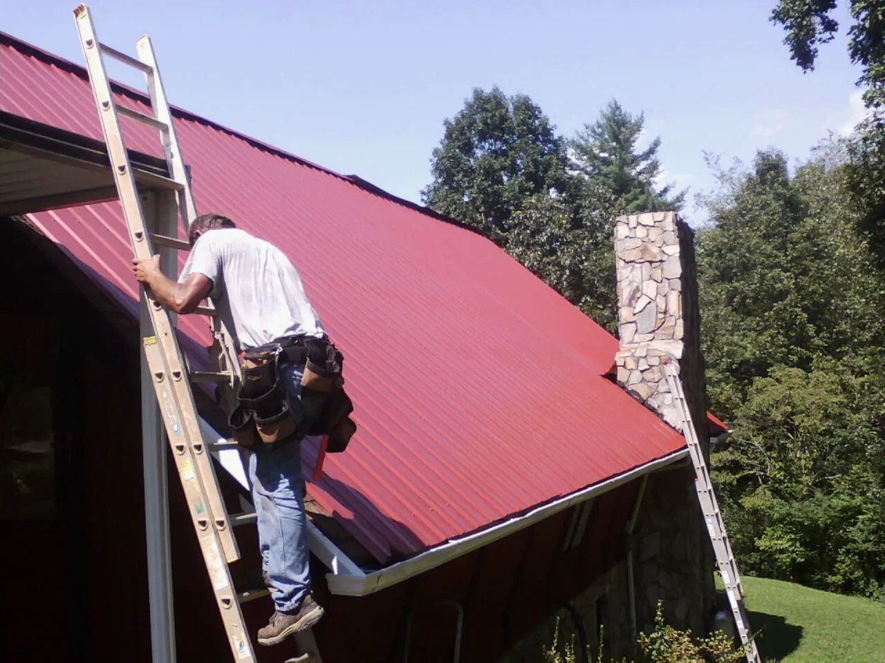 Roofer on a ladder beside a red metal roof and stone chimney — Soffit & Fascia Repair by Roofing Brothers in Armona